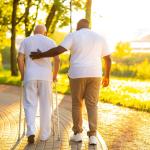 A man in a white shirt helping an elderly man use a walker in a park.