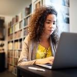 Young adult working on laptop with books on bookshelf in background.