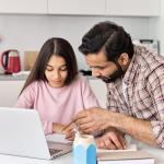 A father and his daughter using a laptop and writing down notes to help with homework.