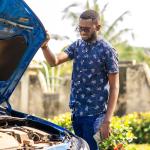 Man with sunglasses stands with the hood of a blue car open, looking at engine, with a nice yard in the background.