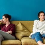 Adult and young adult sitting on opposite sides of yellow couch, looking off into distance in front of blue wall.