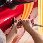 A worker performing dent repairs on the door of a red car.