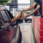 Rear view of drive-thru window worker in red building handing food in brown bag to passenger in passenger seat of black car.