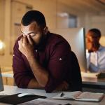 A man in front of a computer at a desk showing a burnout or headache gesture.