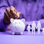 A focus on a person's hand while dropping a coin in a piggy bank with a 3D figure of a 4-member family beside it.