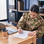 Woman wearing camo military uniform sits at desk doing paperwork.
