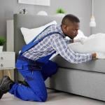 A male exterminator inside a bedroom carefully examining the sheets under a bed.