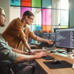 A man working with multiple monitors are engaged in a discussion with his female coworker who points at one of the screens.