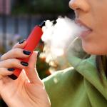 A close up of someone exhaling smoke from vaping, holding vape with black nail polish on fingernails.