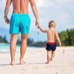 A dad and holding his toddler son's hand while they are walking on a white sand beach.