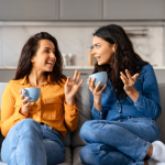 Two young women share stories over tea on a beige couch at home