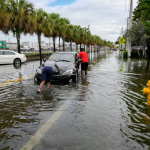 2 people push a car out of one of the flooded streets, after 24 hours of continuous rain in South Florida on June 13, 2024