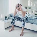 Young adult male sitting on a bed with both hands holding his head seemingly having a headache.
