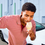 Man eats an ice cream cone in front of a white car in the background.