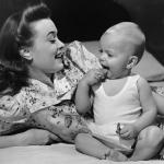 Mother watching her baby eat a cookie, circa 1950s.