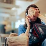 Person on the phone at airport gate, showing signs of frustration covering face with hand while sitting with luggage.