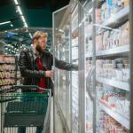 A grocery shopper in a leather jacket pushes a cart through the supermarket and takes food from the fridge.