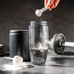 Creatine powder in a measuring scoop being dropped into a clear plastic water bottle, surrounded by workout accessories.