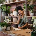 A female owner of a small botanical shop smiling while on the phone taking an order.