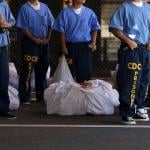 Inmates at San Quentin State Prison shown from the neck down wearing blue uniforms and navy sweat pants that say "CDCR prisoner" wait in line in 2016.