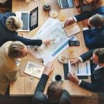 Professionals from an architect business are gathered in a table meeting with food and business documents at the center.