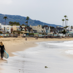 A surfer in a wet suit walking along the beach in Malibu, California carrying a surfboard.