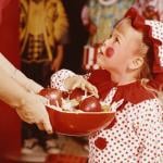 A child in a clown costume choosing a treat from a bowl of apples and candy during Halloween in the 1960s.