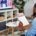 Man watching presidential elections news at home while holding a voting guide.