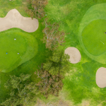 An aerial view of a green golf course with dry patches and trees.