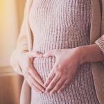 Photo of woman wearing a sweater dress holding hands over belly in a heart shape. 