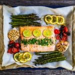 Roasted salmon steak with asparagus, lemon, rosemary, tomatoes, onion, and garlic on a sheet pan.