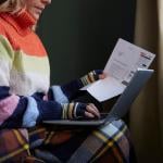 A woman sitting by the radiator is looking at her energy bill and looking up information using a laptop.