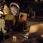 A young man happily opening Christmas presents while in a video call with someone on a laptop.