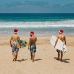 Three people walking on the beach holding their surfboards and wearing Santa hats.