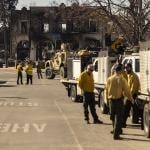 A charred building is seen in the distance as private firefighting company employees, hired to protect Rick Caruso's Palisades Village mall from the Palisades Fire, gather near their vehicles.