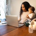 An African-American mother holds her baby daughter while taking notes and using a laptop.