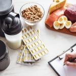 A set of food supplements such as protein powder and fish oil supplements on a table with fresh food while a person writes a diet plan.