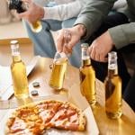 A group of three male friends consuming beer and eating pizza while playing video games.