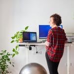Woman working from home with an adjustable desk standing next to a fitness ball.