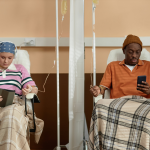 Two people sitting side-by-side and scrolling on their phones while getting chemotherapy treatment in clinic