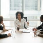 Black woman at head of table leads discussion with colleagues on each side of table.