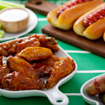 A table with glazed chicken wings, hot dogs, celery sticks and dip