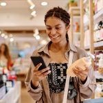 A smiling woman in a store, holding a bottle with a pump and looking at her phone. 