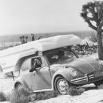 A passenger smiles inside a Volkswagen Beetle traveling with a Kangaroo camper through the desert.