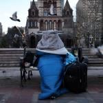 Bundled in blankets and a sleeping bag, a homeless person sits on a bench in Copley Square in Boston. 