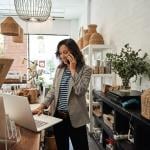 A small business owner looking at her laptop and talking on the phone behind the counter in her boutique.