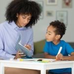 Black adult helping child learn and write, looking at a book.