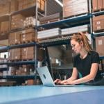 A woman managing logistics and using a laptop in a warehouse.