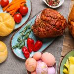 Easter dishes such as honey ham, baked buns, potatoes and colorful easter eggs on a wooden table.