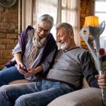 An elder couple where the wife shows her phone to her husband who is holding on to a pair of crutches while sitting on a sofa.